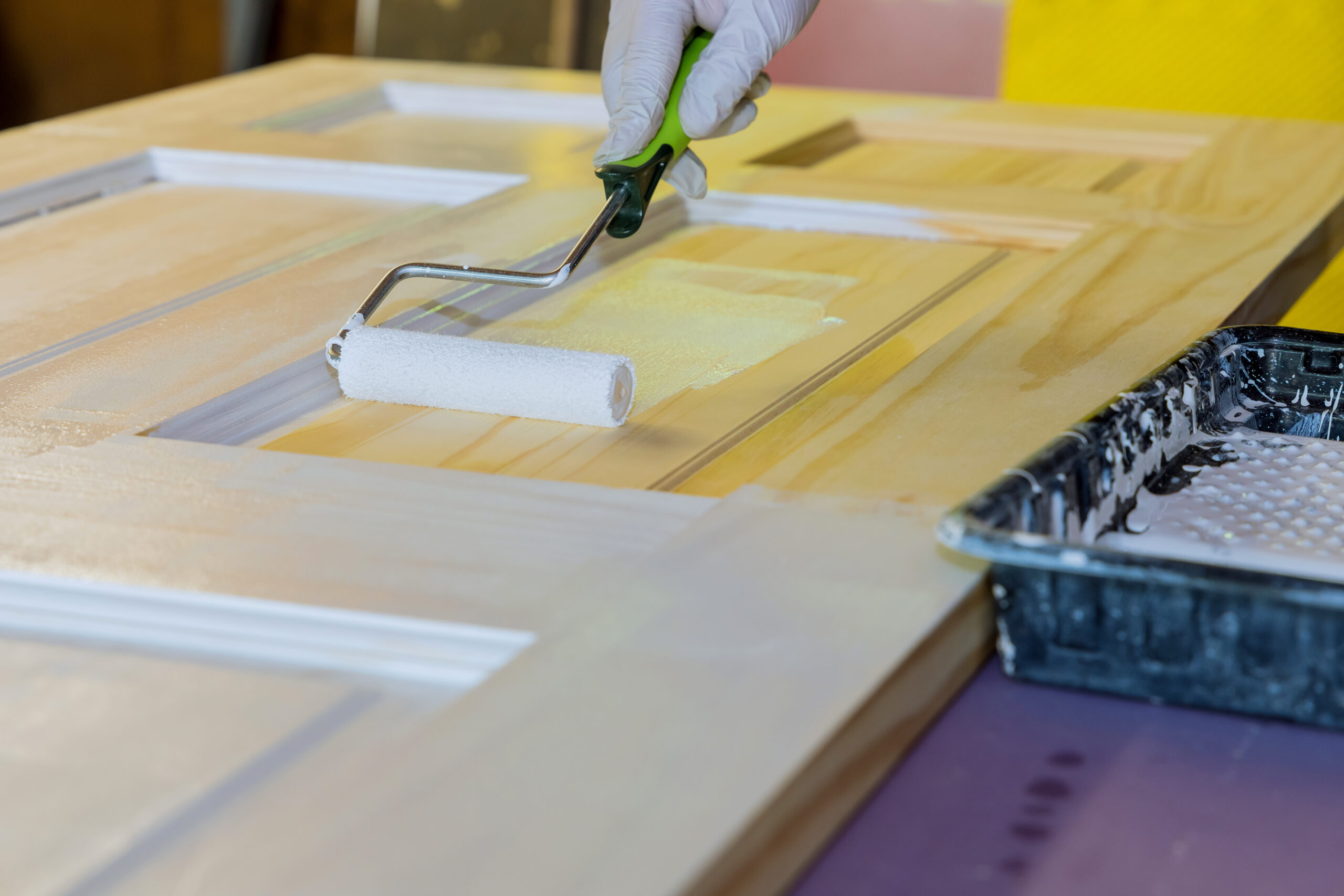 Close-up of a gloved hand using a paint roller to apply white paint to a paneled wooden door, with a black paint tray nearby on the work surface.