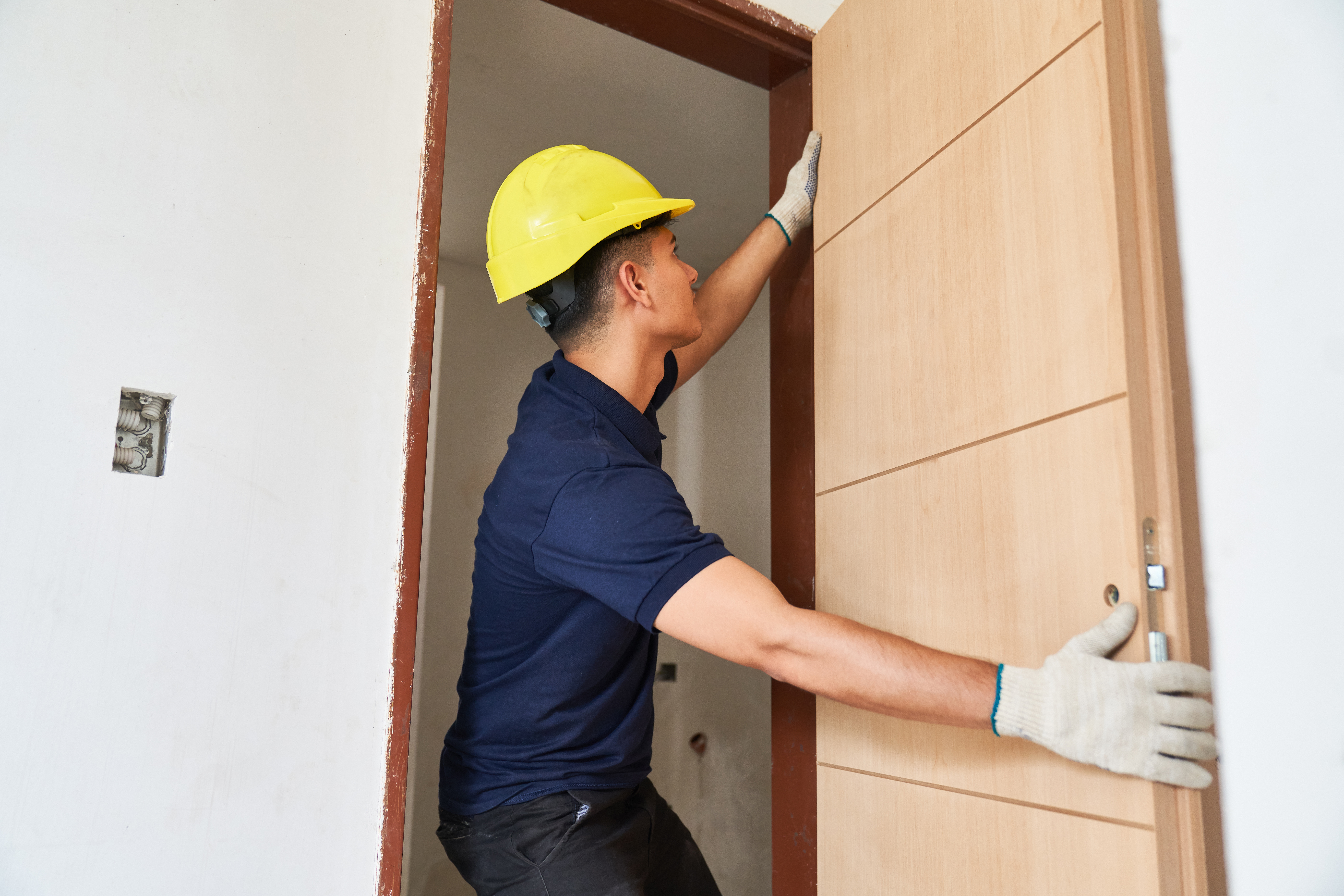 A worker in a yellow hard hat installing a modern wooden door into a doorway, carefully aligning it within the frame.