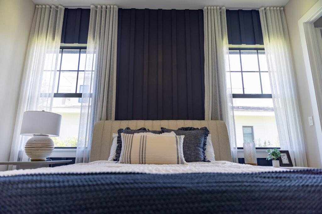 Modern bedroom featuring a dark vertical accent wall behind a light upholstered bed, framed by two windows with sheer white curtains and soft natural light.
