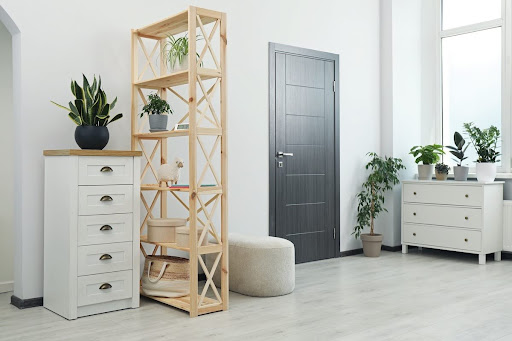 Bright modern room with white walls, light wood floors, a dark gray interior door, white dressers, a light wood shelving unit, and several potted plants near a large window.